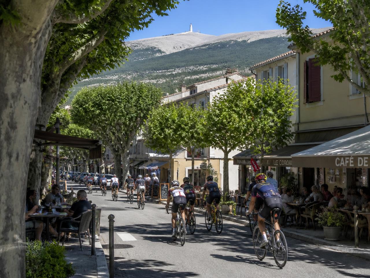 Le cours ombragé de Malaucène avec ses terrasses et des cyclistes, le sommet du Mont Ventoux en arrière-plan
